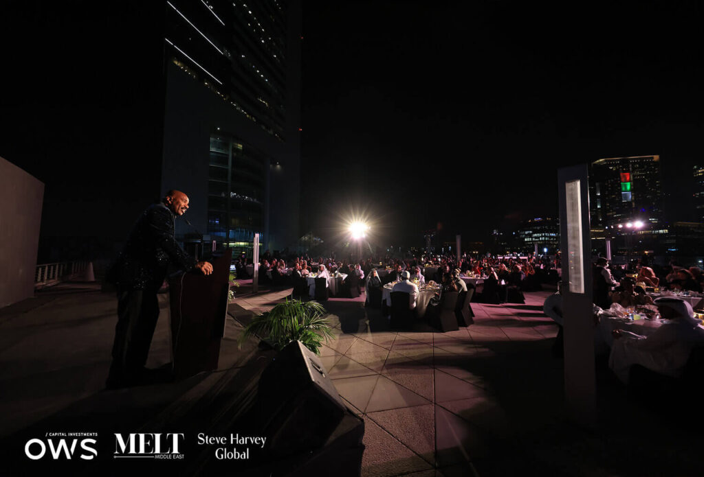 Steve Harvey at podium with guests at their tables in the background
