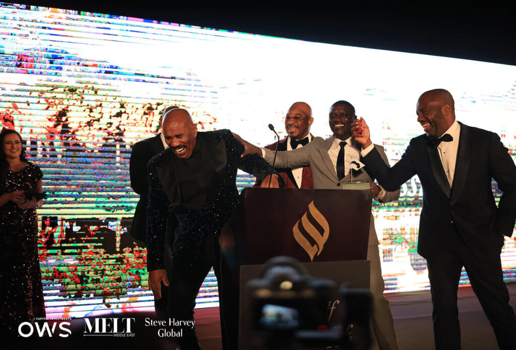 Steve Harvey laughing at podium with other guests on the stage